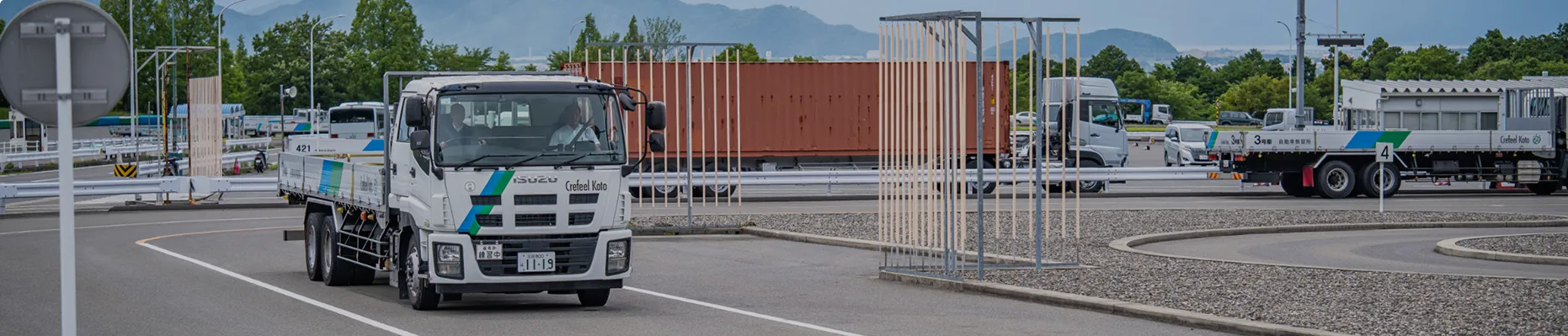 教習所コースを走行する大型トラックと、運転席・助手席に乗る二人の男性。