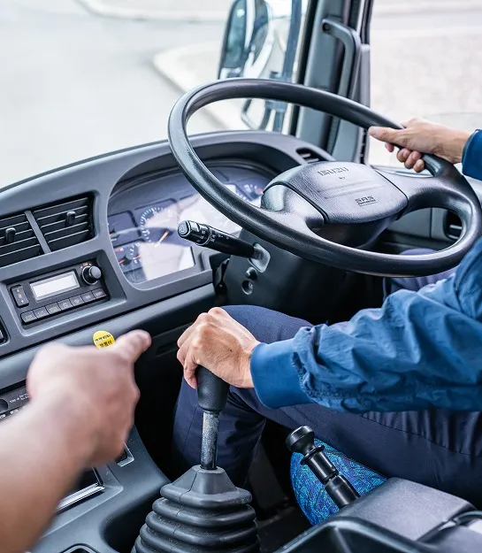 大型トラックの運転席で、シフトノブを操作する教習生の手元と、それを指差して指導する指導員の手。