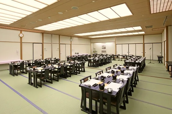 広々とした和室の大宴会場。畳の上に黒いテーブルと椅子席が多数並べられており、会席料理がセッティングされています。奥には床の間やマイクが用意された、落ち着いた和の空間です。