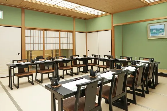 落ち着いた和室の小宴会場。畳の部屋に黒いテーブルと椅子席がコの字型に配置され、お食事がセッティングされています。緑色の壁や障子に囲まれた、少人数で和やかに会話ができる空間です。