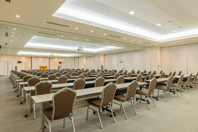 明るく広々とした研修室。手前に講師用の演台があり、受講者席には白い1人掛けのデスクと青い椅子が前方に向かって整然と並んでいます。正面の壁には大きなホワイトボード、天井にはプロジェクターが設置されており、左側には大きな窓があります。