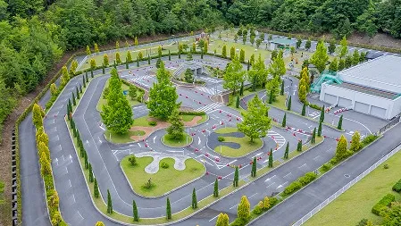 上空から撮影した施設全景。木々に囲まれた広い敷地の中に、白線や横断歩道が引かれたうねるコース、芝生エリア、右奥には白い建物が見える。