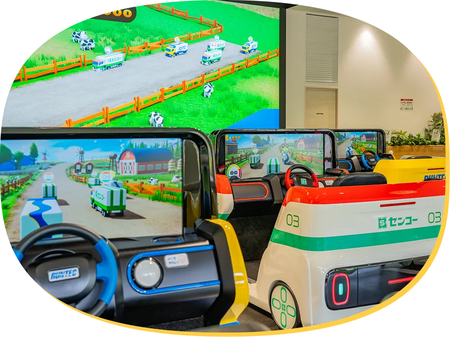 牧場のようなコースをCGのトラックが走るゲーム画面が、運転席前方のモニターと正面の大画面に映し出されている様子。