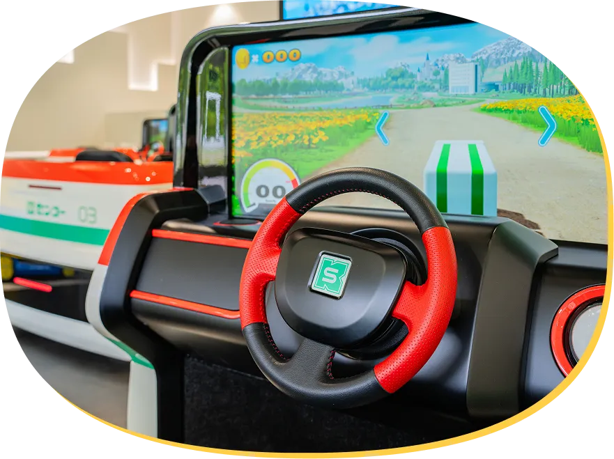 赤と黒の本格的なハンドルを備えた運転席のアップ。前方のモニターには、お花畑や緑の風景が広がるドライブコースが映し出されている様子。