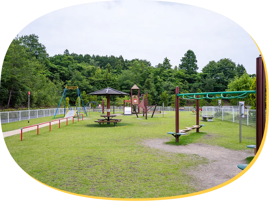 手前に緑色のうんていやステップ遊具があり、奥には東屋やターザンロープ、複合遊具がバランスよく配置された広々とした芝生エリア。