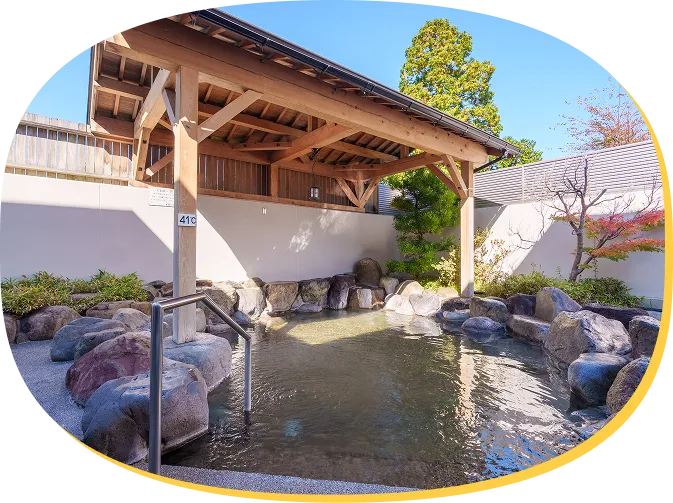 至福の湯の露天風呂。立派な木造の屋根に守られた岩風呂で、周囲には緑の植栽や石畳がある。手すり付きの階段で湯船に入る。