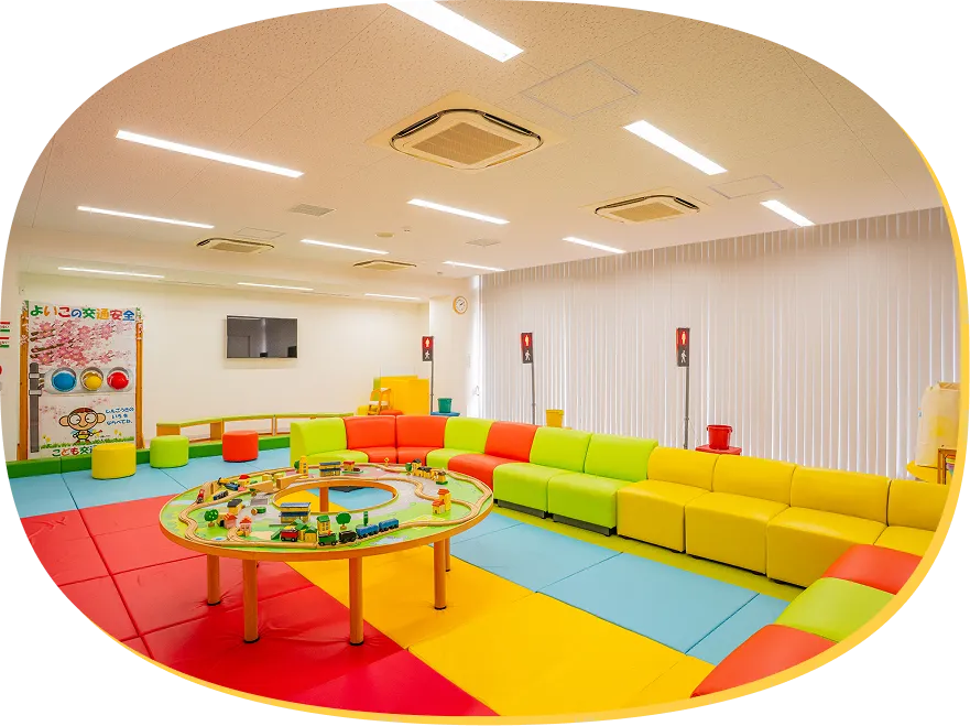 赤、黄、緑などのカラフルなソフトマットが敷かれ、壁沿いに低いソファが並ぶキッズルーム。部屋の中央には、木製レールの汽車のおもちゃが置かれた円形テーブルが設置されている様子。
