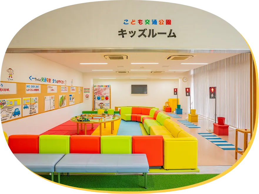 上部に「こども交通公園 キッズルーム」と掲示された入り口からの全景。左の壁には交通安全のお約束が展示され、奥にはカラフルなソファと体験用信号機が並んでいる様子。