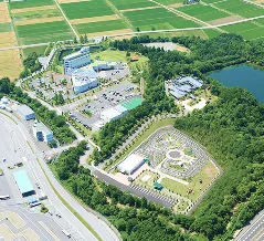 幹線道路に隣接し、広い駐車場を備えた緑豊かな施設全体の空撮写真。