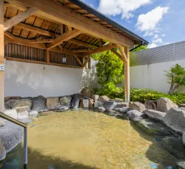 大きな岩と木造の屋根に囲まれた、青空が覗く開放的な大浴場の露天風呂。