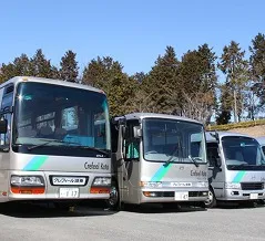 青空のもとに並ぶ、施設ロゴが入ったシルバーの送迎用シャトルバス。